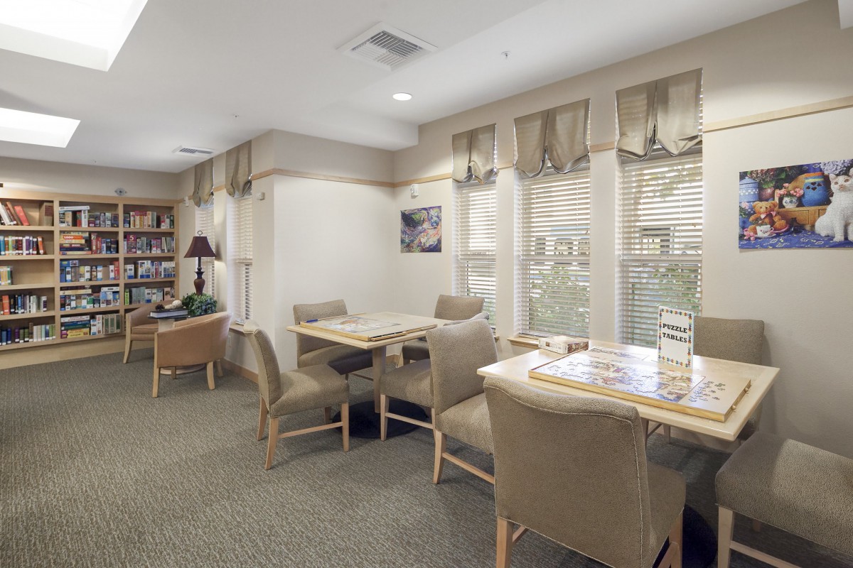 A cozy waiting room with tables, chairs, bookshelves, and large windows with curtains.