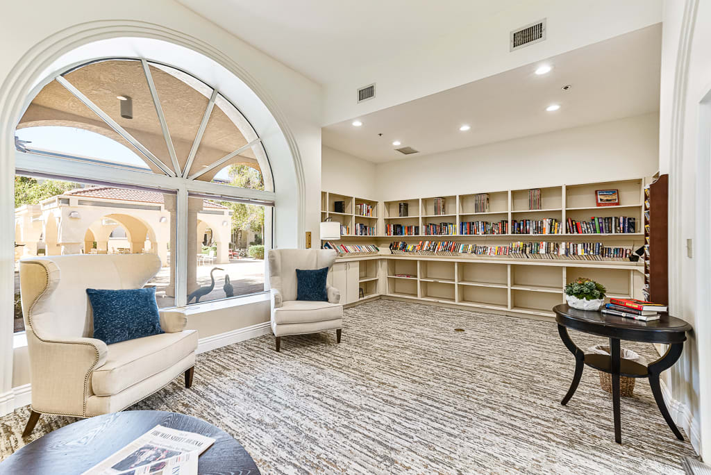 A cozy library reading room with bookshelves, comfortable chairs, and large windows.