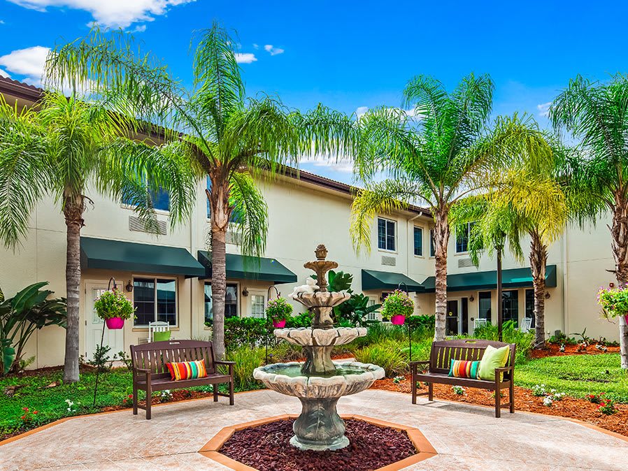 A vibrant courtyard featuring a central fountain, surrounded by palm trees and benches, with buildings in the background.