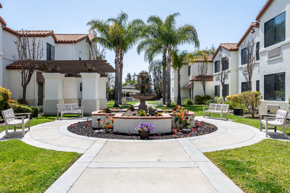A picturesque residential area with apartments flanking a central walkway and fountain, surrounded by lush palm trees.
