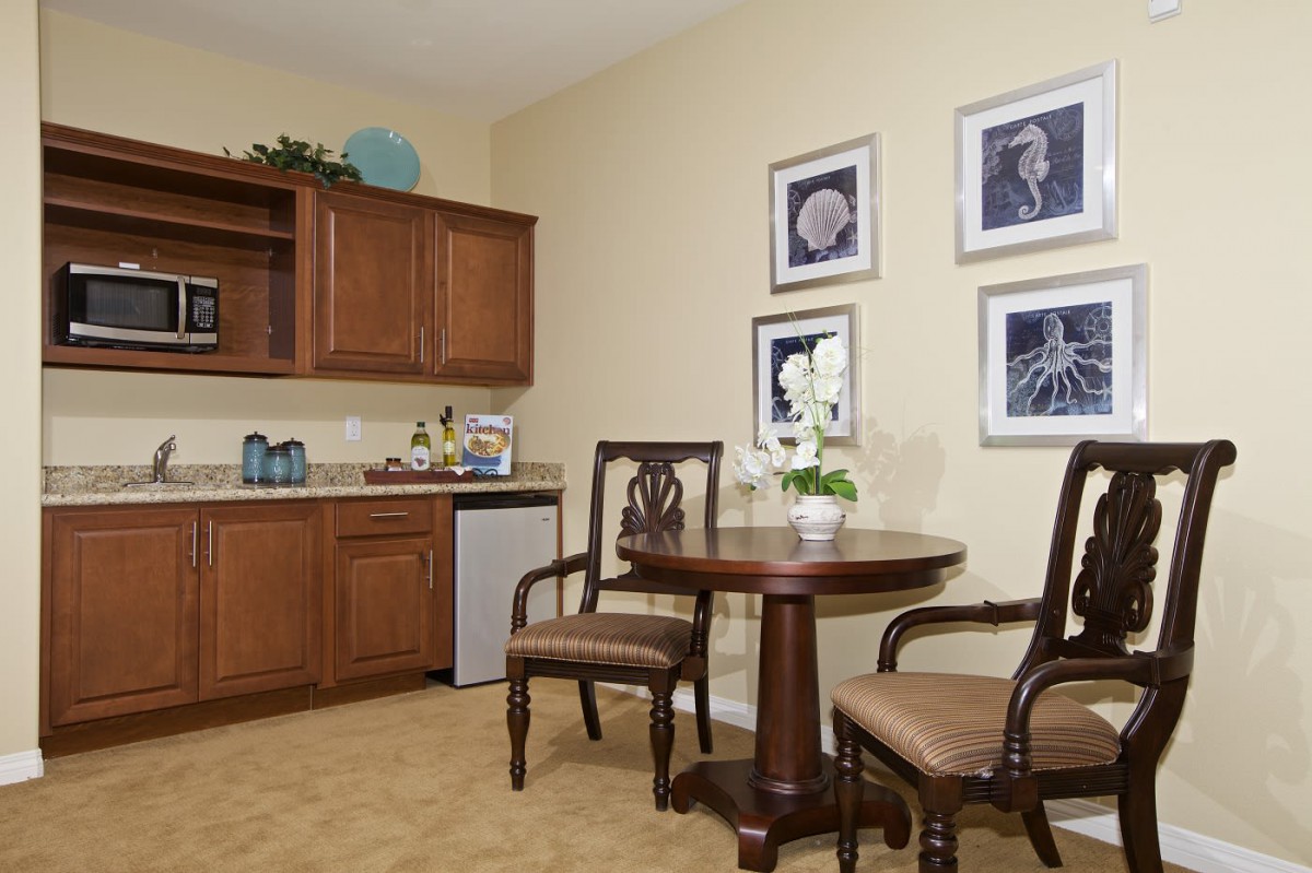 A cozy kitchenette with wooden cabinets, a small table and chairs, decorated with artwork and plants.