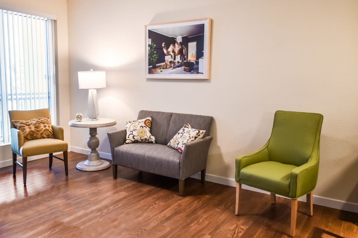 A cozy living room area featuring a small couch, a green chair, and a side table with a lamp, set against wood flooring and decorated with wall art.