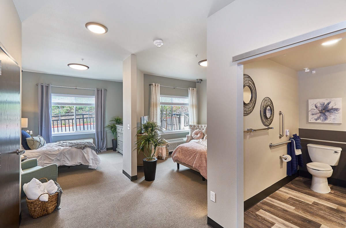 A bright and spacious bedroom with two beds, decorated with plants and ample natural light from large windows.