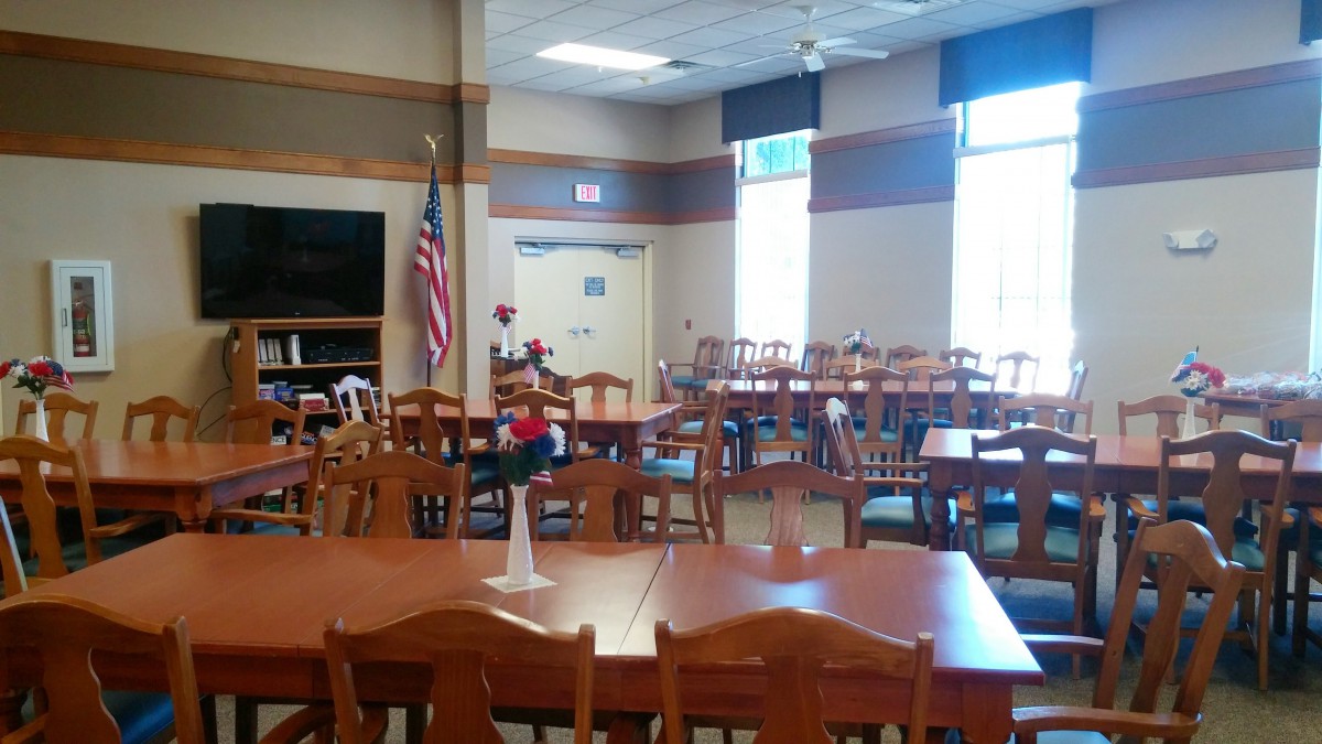 An empty dining room with wooden tables and chairs, featuring an American flag and large windows.