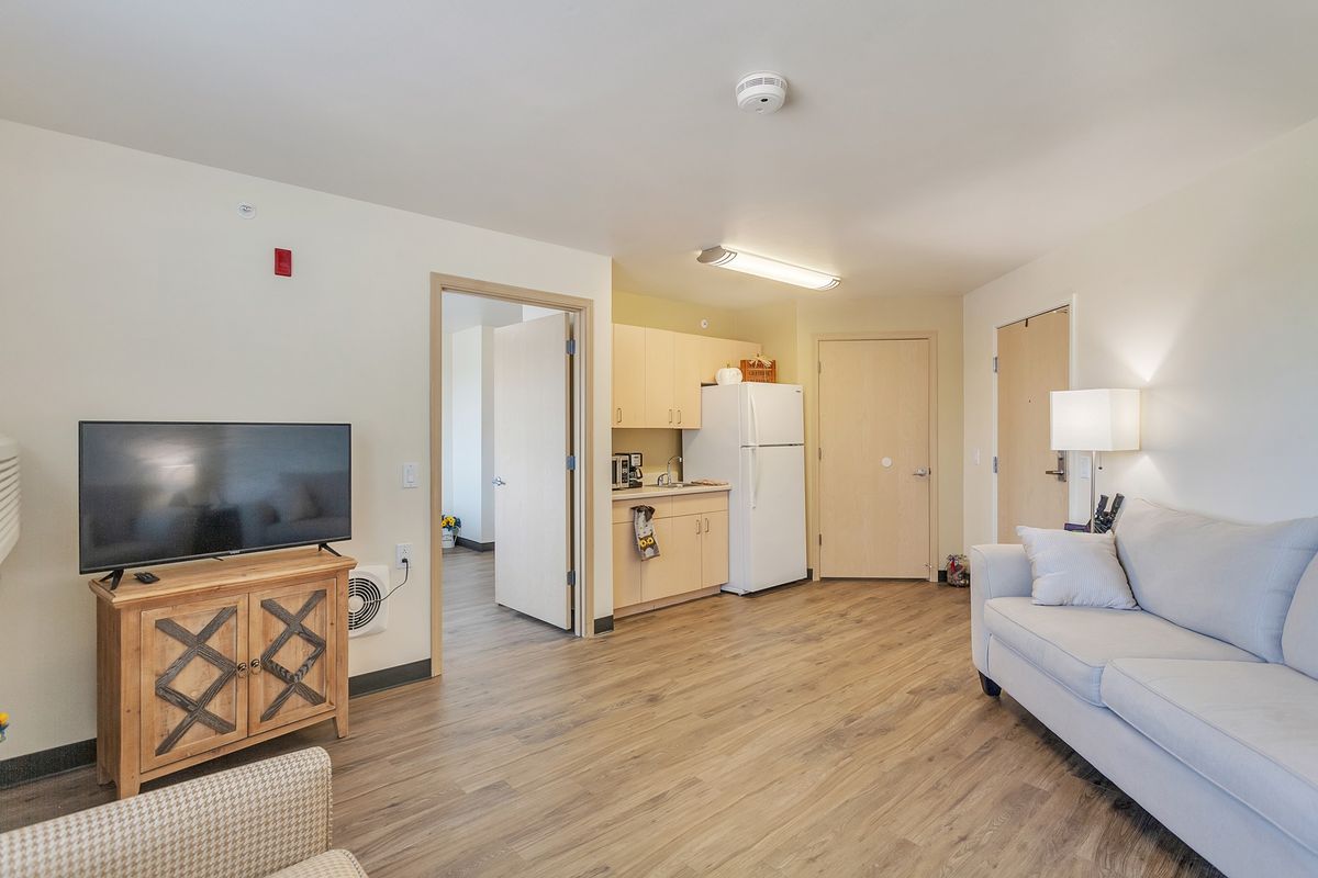 A modern apartment living area with an open kitchen, featuring a sofa, TV, and refrigerator.