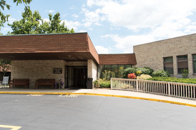 The image shows the entrance of a building with landscaped surroundings and a parking lot under a partly cloudy sky.