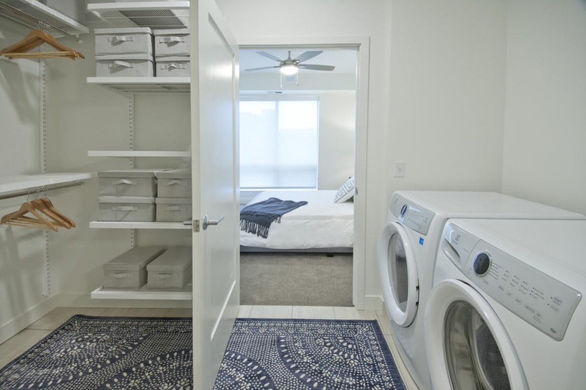 View from a laundry room into a bedroom, showcasing appliances and shelves in a modern home setting.