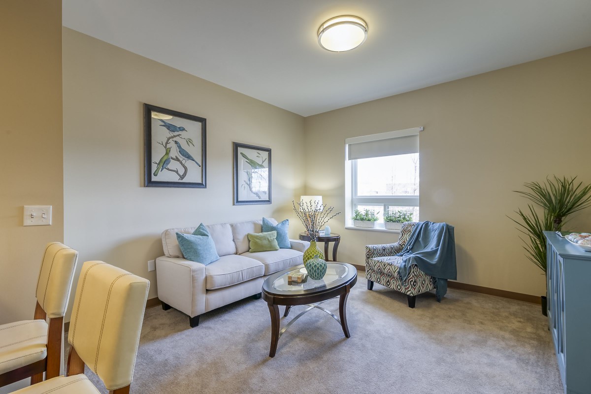 A cozy living room with a sofa, armchair, and coffee table, decorated with art and featuring a window for natural light.