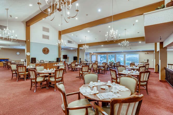 A spacious and elegant dining room with multiple round tables and wooden chairs, illuminated by chandeliers.