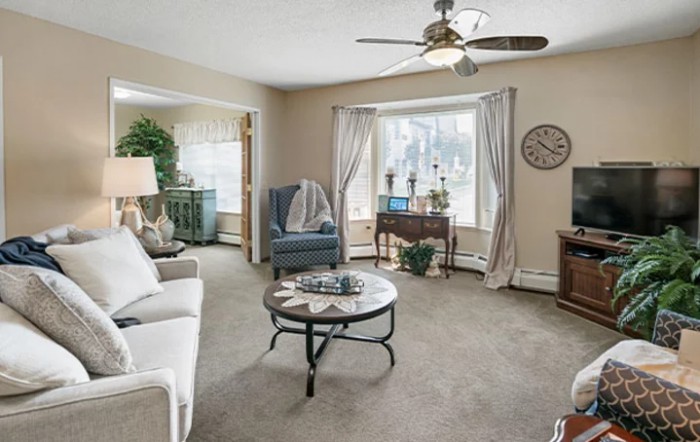 A cozy living room with a sofa, armchair, and coffee table, featuring large windows with curtains and a ceiling fan.