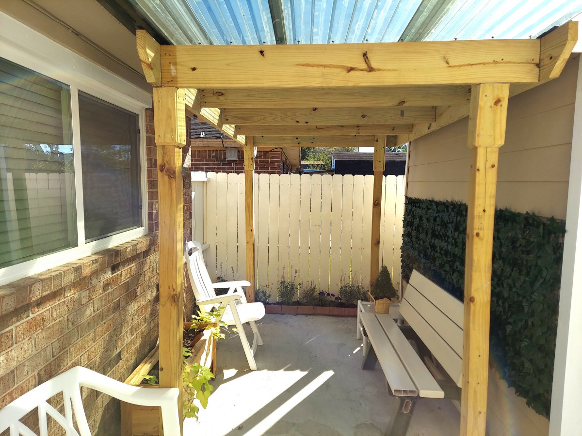 A cozy patio with a wooden pergola, featuring outdoor seating including a bench and chairs.