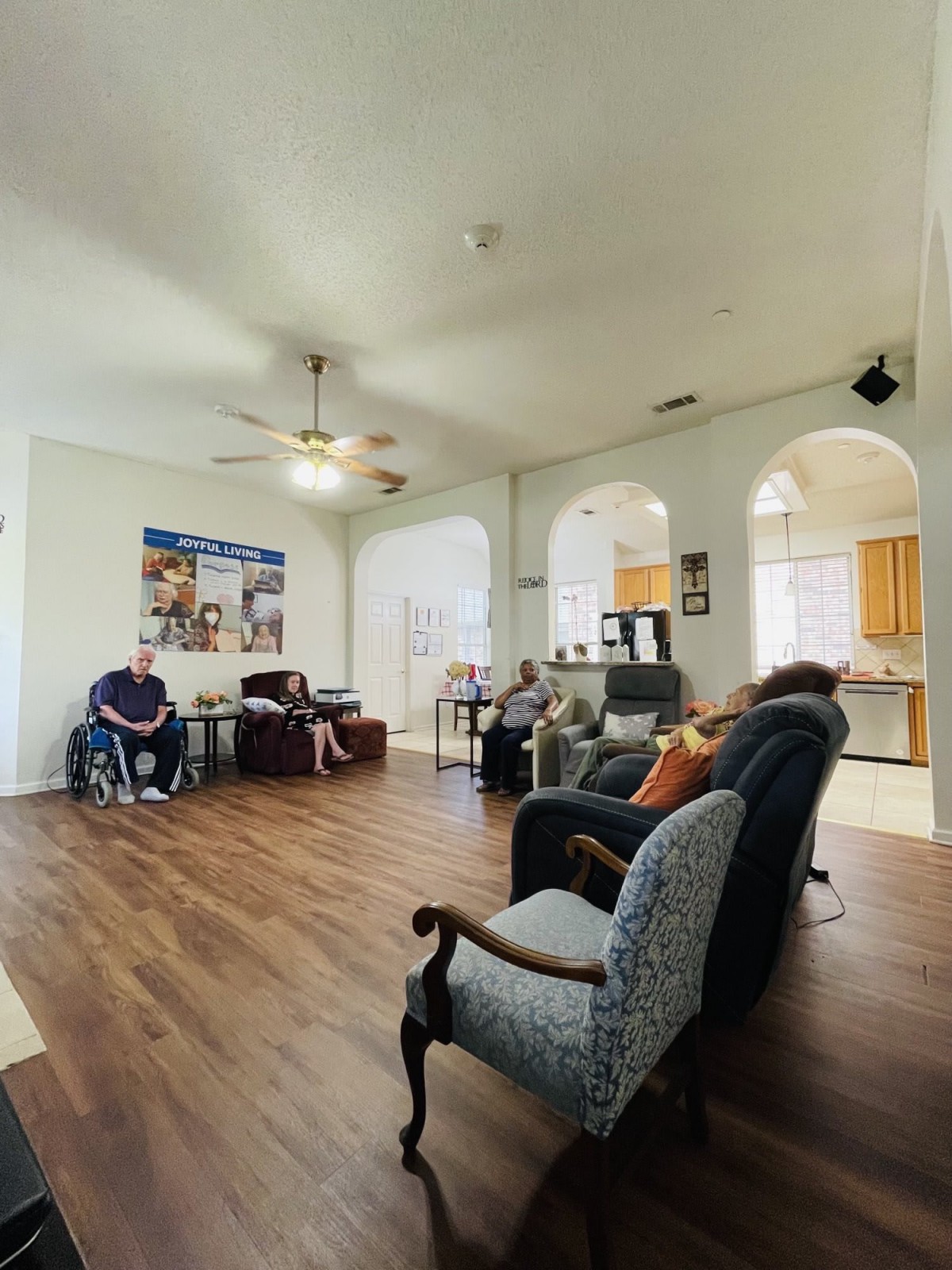 A spacious living room with a ceiling fan, hardwood floor, and various seating options, connected to an open kitchen through arched doorways.
