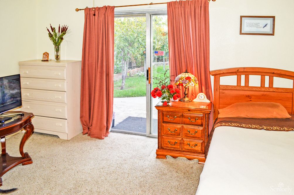 A cozy bedroom with wooden furniture, pink curtains, and a view of the garden through a sliding glass door.