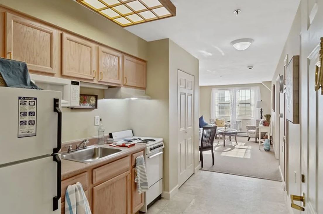 A cozy apartment kitchen with wooden cabinets, adjacent to a bright living area.