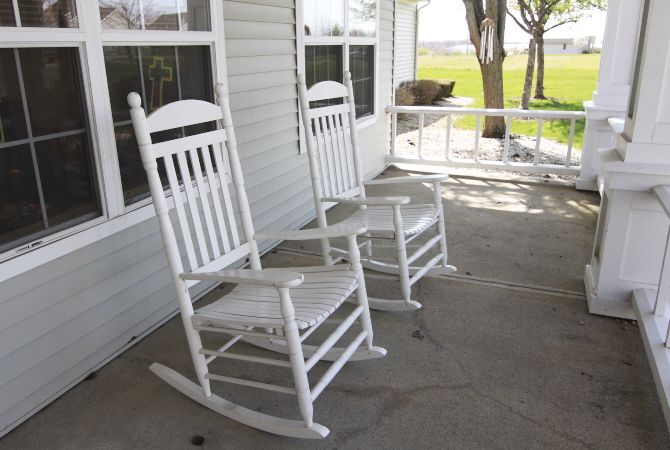 Two white rocking chairs sit on a porch, overlooking a green lawn.