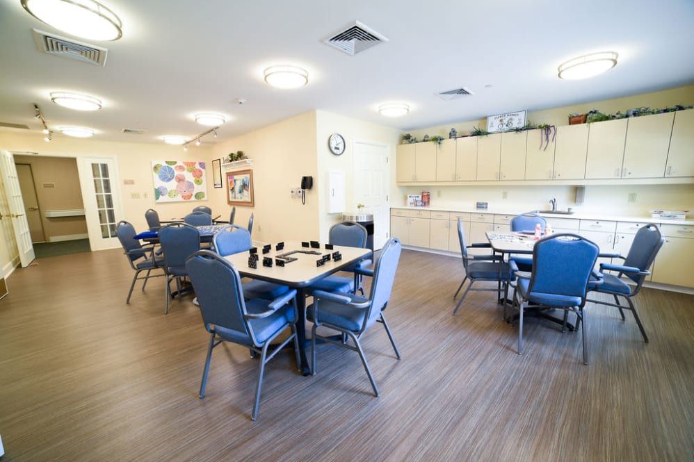 A bright community room with several tables and chairs, featuring a small kitchen area.