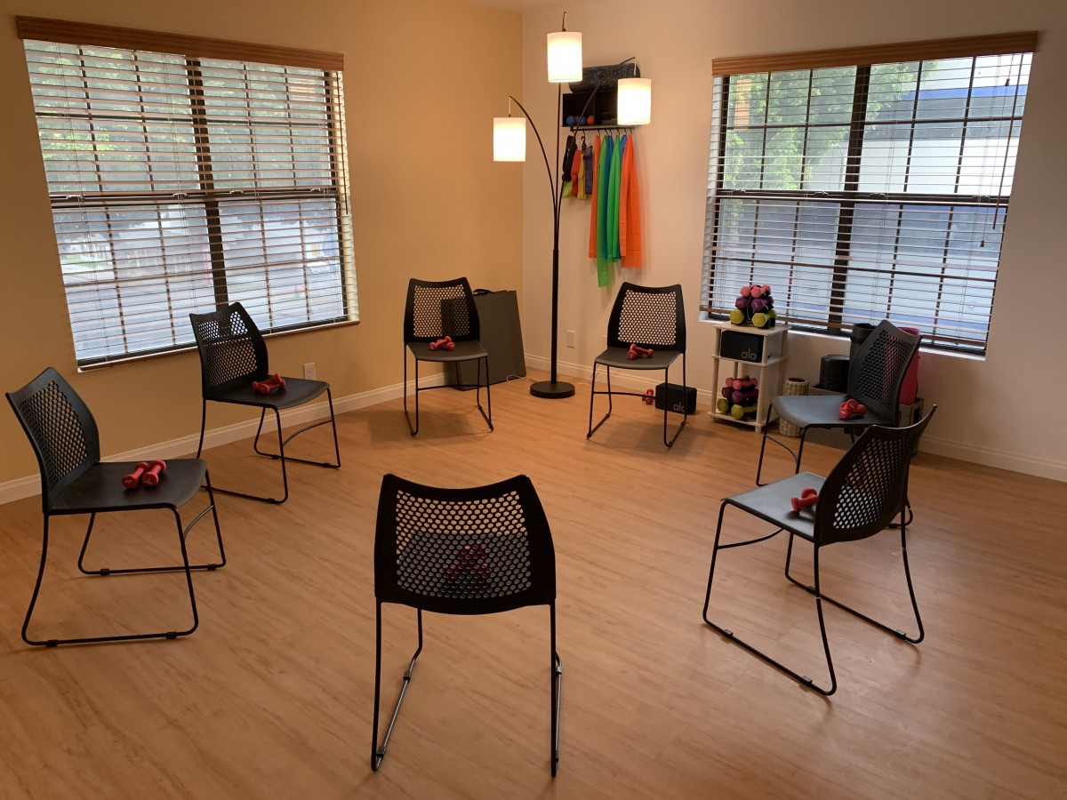 A therapy or meeting room arranged with chairs in a circle, featuring large windows and wooden flooring.