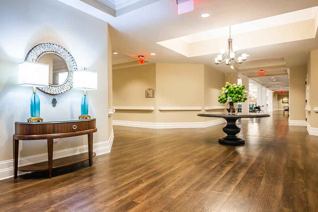 A spacious and elegantly decorated lobby with wood flooring, featuring a side table with a decorative mirror and a central round table adorned with flowers.