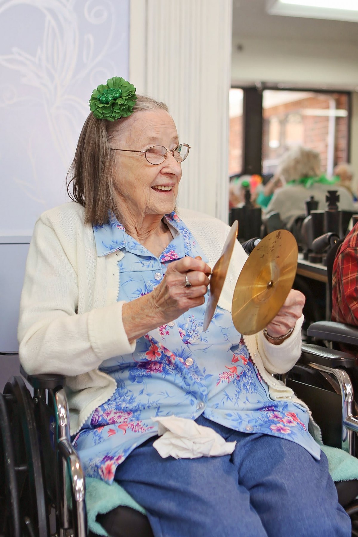 An elderly woman wearing a blue floral shirt and cardigan is seated in a wheelchair, smiling as she holds a pair of cymbals.