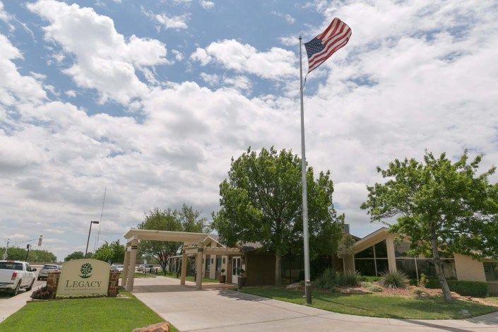 An American flag flies high near a building surrounded by trees under a partly cloudy sky.