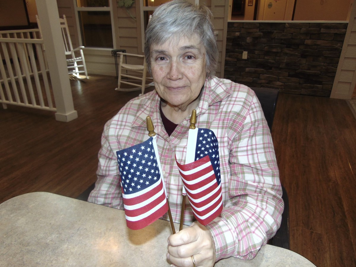 An elderly woman sitting indoors holding two small American flags.