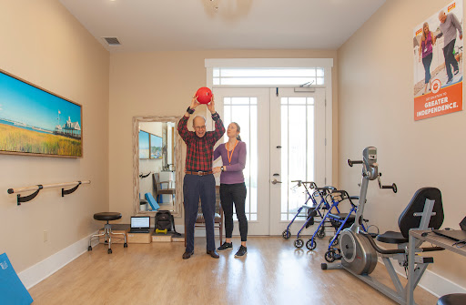 A fitness instructor guides a senior individual in a rehabilitation exercise using a medicine ball in a bright indoor setting.