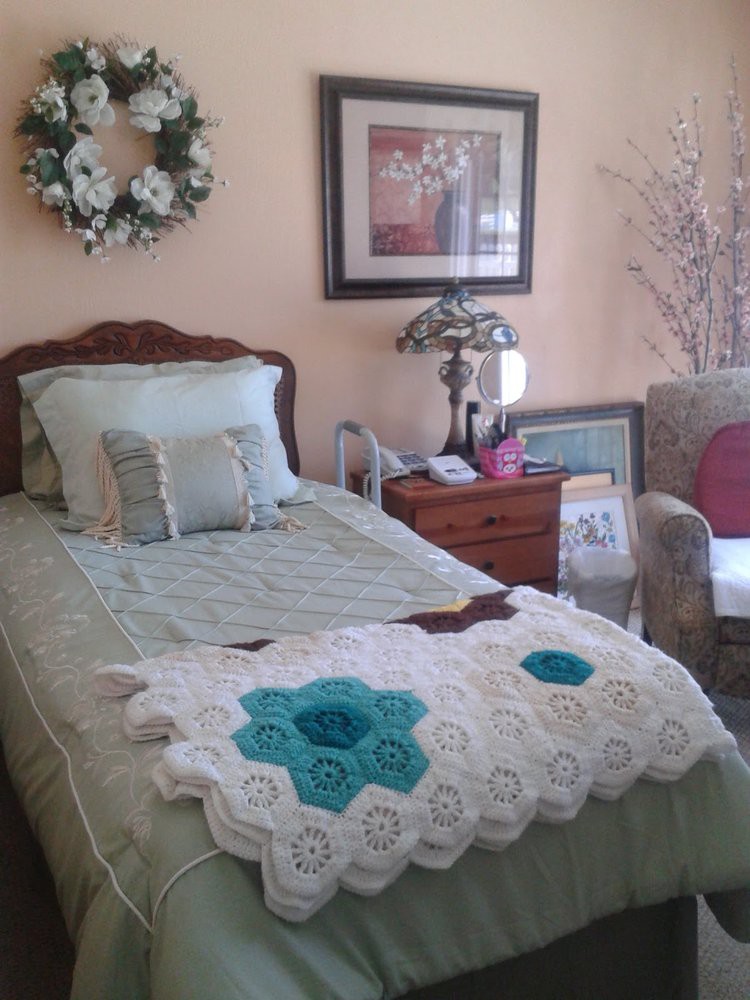 A cozy bedroom with a single bed adorned with a crochet blanket and decorative pillows, accompanied by a nightstand, lamp, and framed art on the wall.