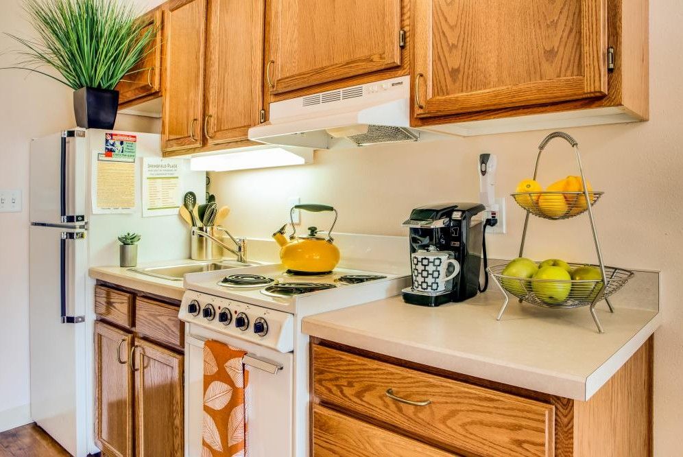 A cozy kitchen with wooden cabinets, a stove, and countertop appliances including a teapot and coffee maker, alongside a fruit basket.