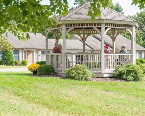 A serene garden scene featuring a wooden gazebo with people sitting inside, surrounded by a lush green lawn and trees.
