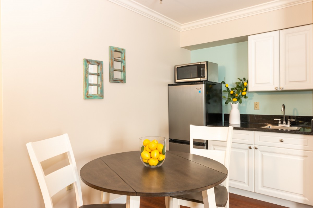 A cozy kitchen nook featuring a wooden dining table with lemons, surrounded by white chairs, and a kitchenette with modern appliances.