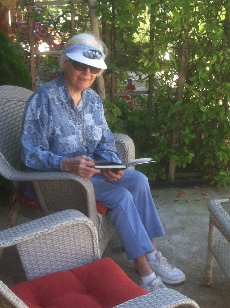 A person sitting outdoors in a chair, wearing a hat and glasses, reading.