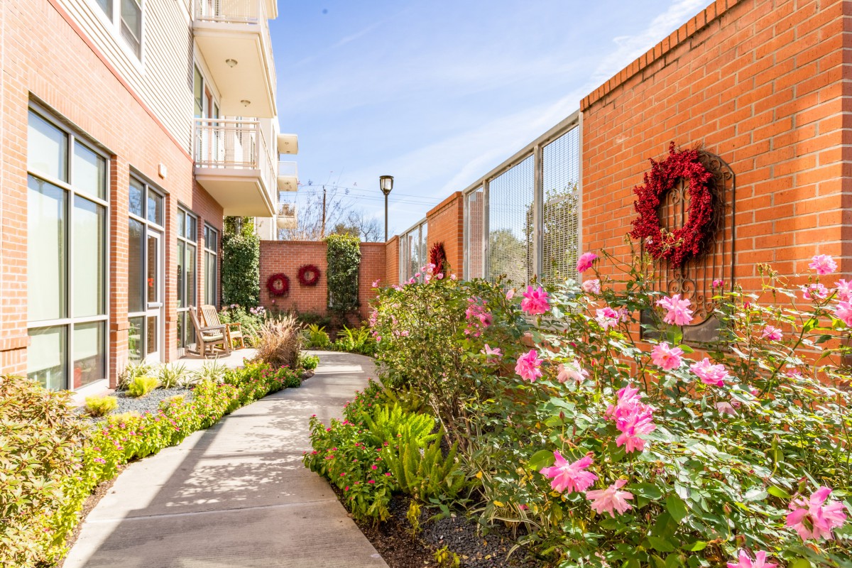 A landscaped garden with blooming flowers and a pathway alongside brick walls adorned with wreaths, under a clear sunny sky.