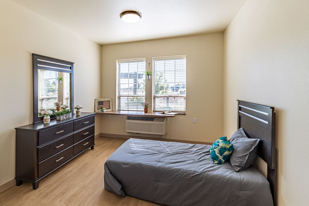 A minimalist bedroom with a single bed and dresser, featuring natural light from two large windows.