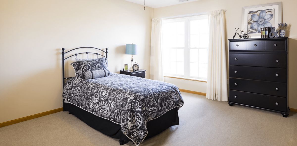 A cozy bedroom with a single bed dressed in patterned bedding, accompanied by a nightstand and lamp near a large window.