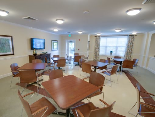 A community dining area with multiple tables and chairs, featuring a television on the wall and large windows with curtains.