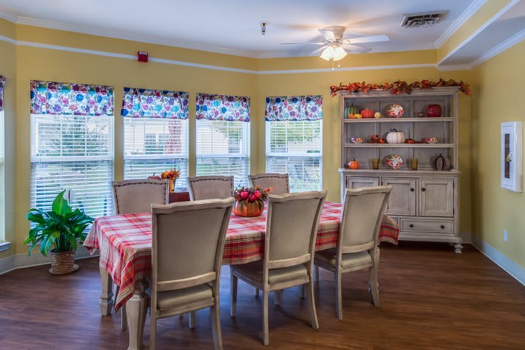 A cozy dining room with a large table and chairs, surrounded by windows and decorated shelves.