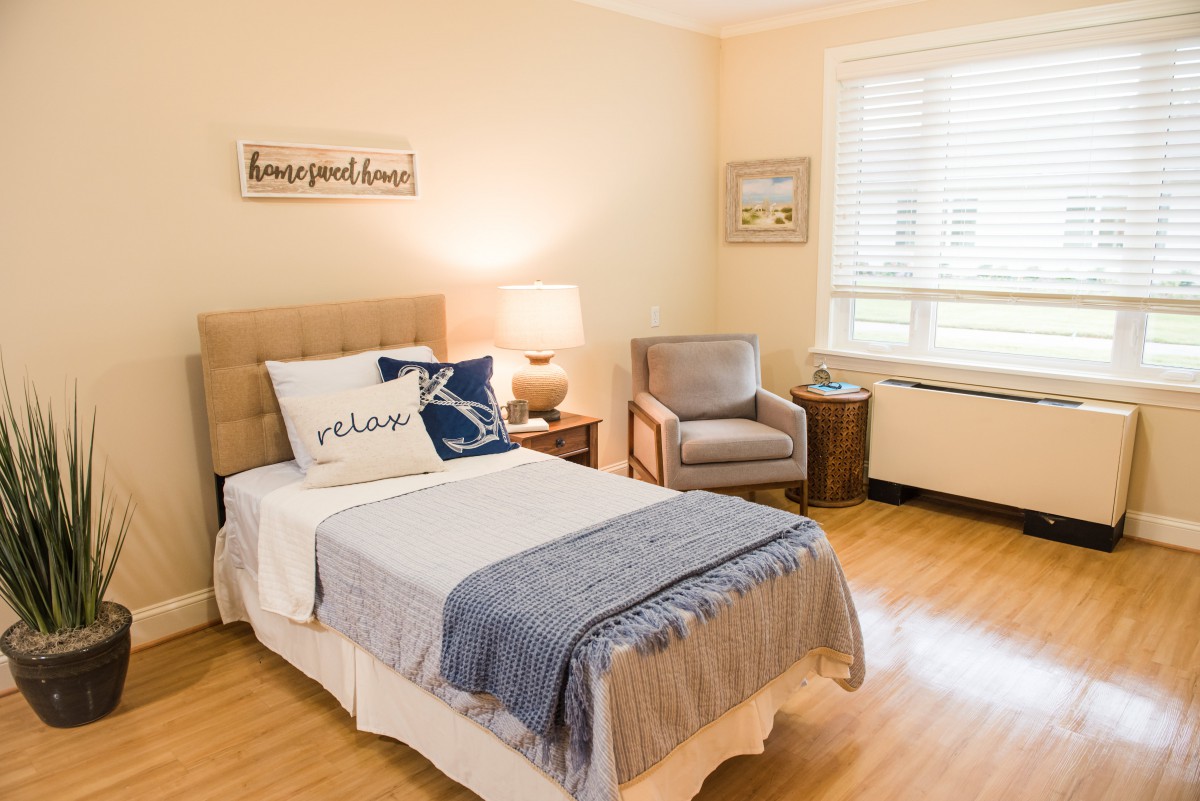 A cozy bedroom with a single bed, decorative pillows, a lamp on a side table, and an armchair by the window.
