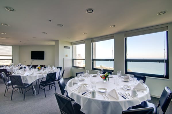 A well-lit dining room with round tables set for an event, offering a stunning ocean view through large windows.