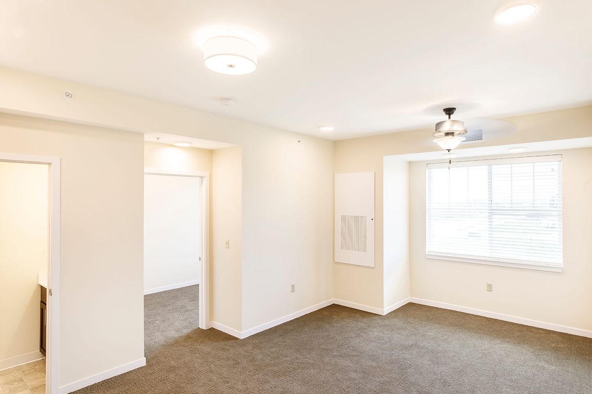 An empty living room with carpet flooring, a ceiling fan, and a large window.