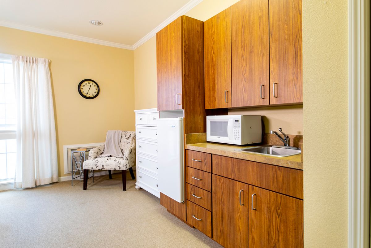 A cozy kitchen corner with wooden cabinets and a microwave on the countertop, featuring a decorative chair and clock against beige walls.