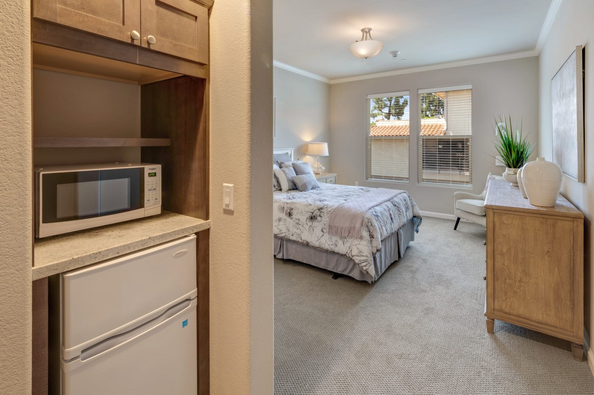 A cozy bedroom with a small kitchenette area, featuring a bed, bedside lamp, microwave, and mini fridge.