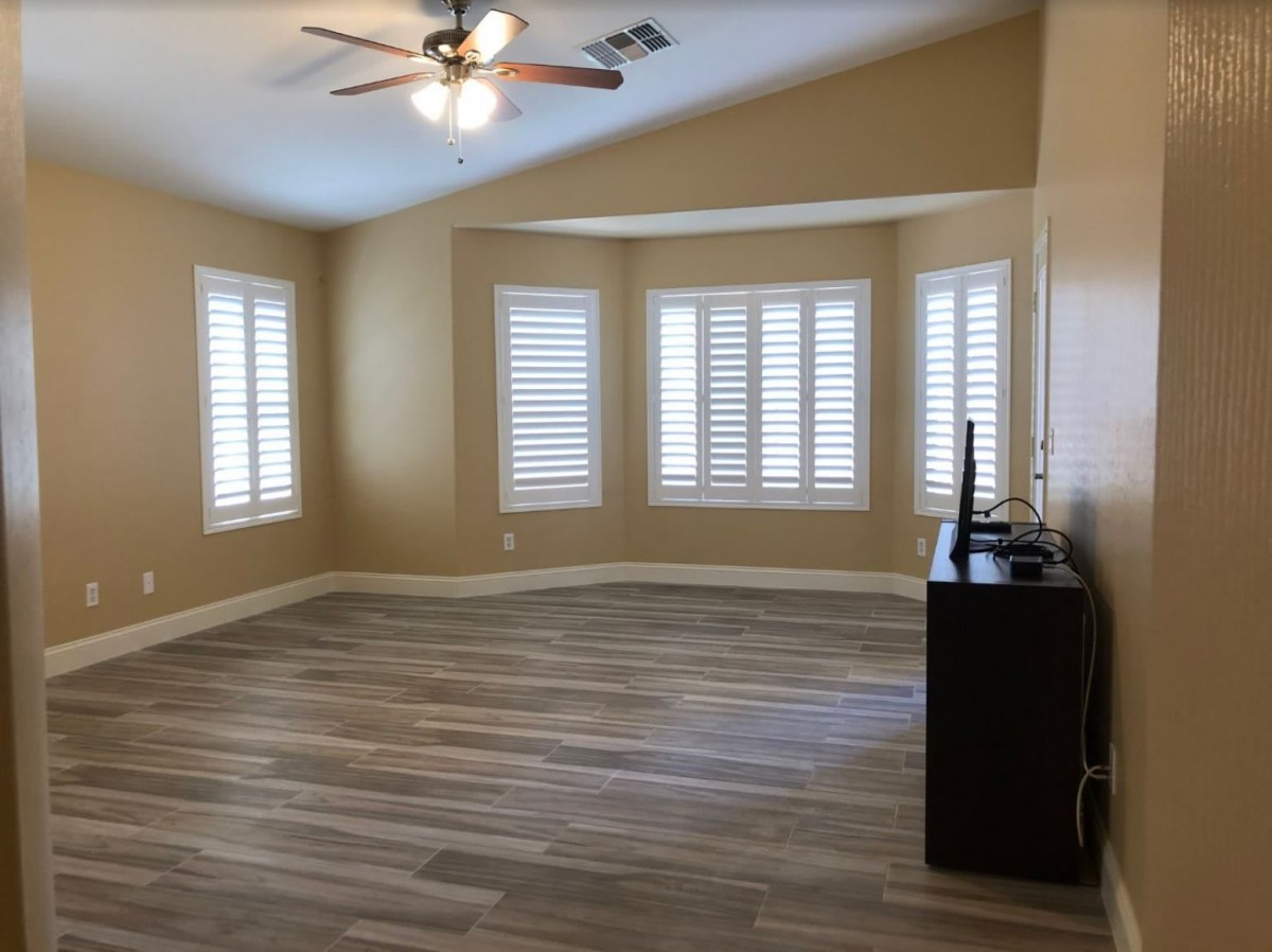 An empty living room with wood flooring and a ceiling fan, featuring multiple windows with blinds.