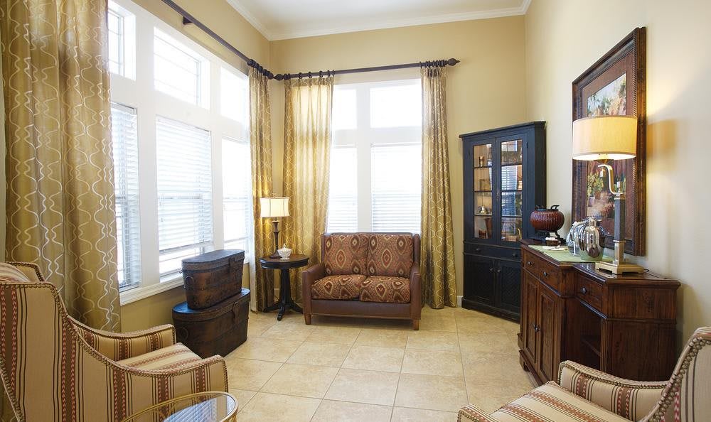 A cozy living room with ample natural light, featuring an armchair, cabinet, and elegant curtains.