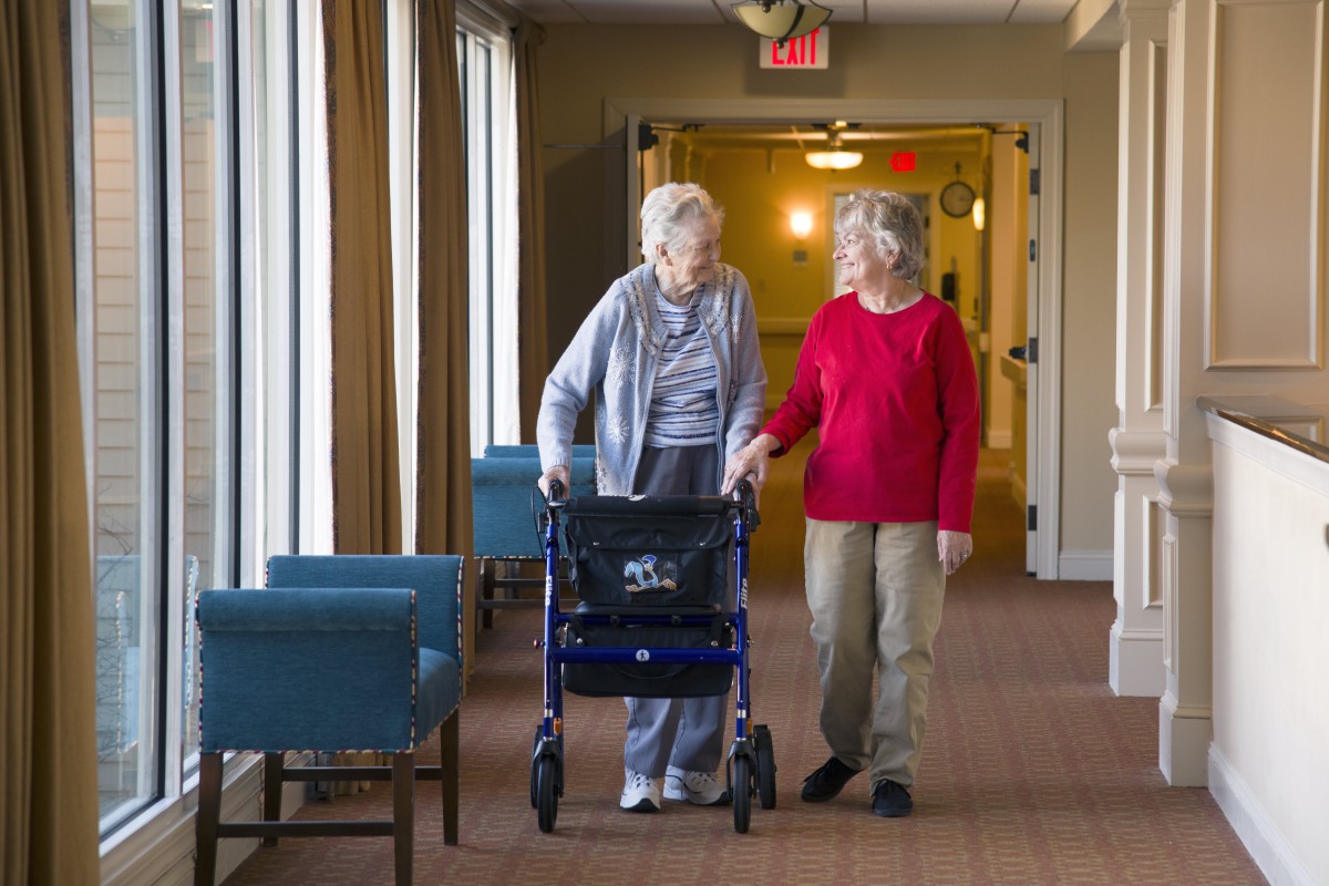 Two elderly individuals walking in a hallway, one using a walker and the other providing support.