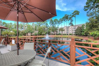 A serene patio with outdoor seating overlooking a lake and fountain surrounded by trees and apartments.