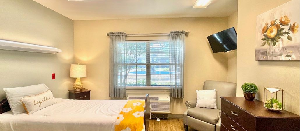 A cozy bedroom with a large window, bed, armchair, and wall-mounted TV.