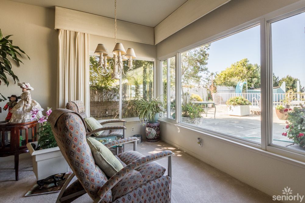 A cozy living room with large windows overlooking a garden, featuring comfortable chairs and indoor plants.