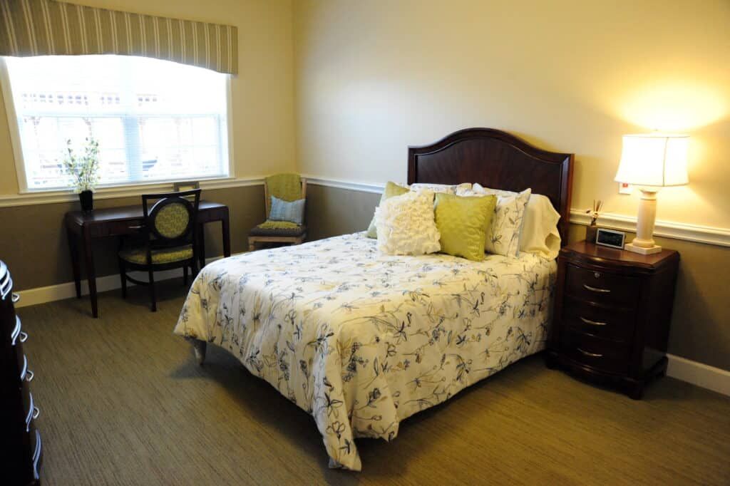 A cozy bedroom with a neatly made bed, adorned with decorative pillows, a bedside lamp, and a window letting in natural light.