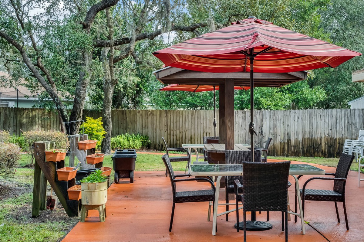A cozy backyard patio with tables and chairs under umbrellas, surrounded by trees and a wooden fence.
