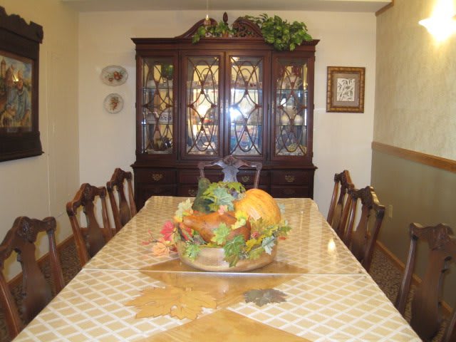 A dining room with a long table and chairs, featuring a decorative centerpiece, with an elegant cabinet in the background.